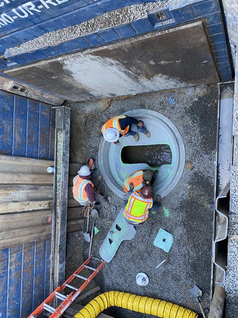 Crew setting a precast structure at the bottom of a shored trench, with forced-air ventilation ducting run down to them.