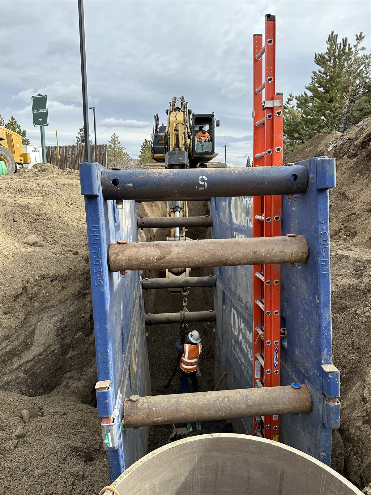 A shored trench with hydraulic shields and struts, ladder egress in place, an excavator lowering pipe to the crew below.