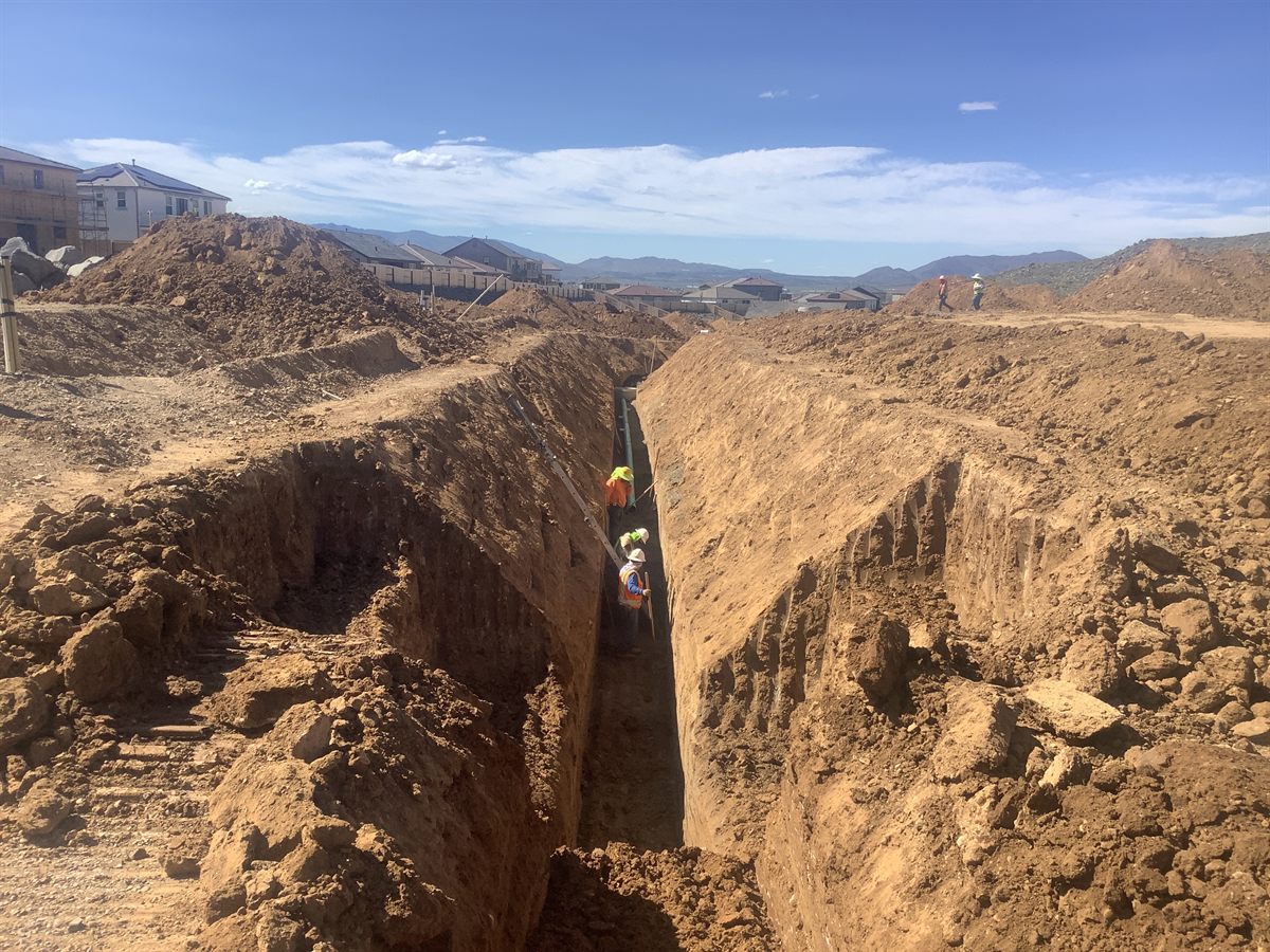 A deep utility trench cut through hillside ground with crew working at the bottom.