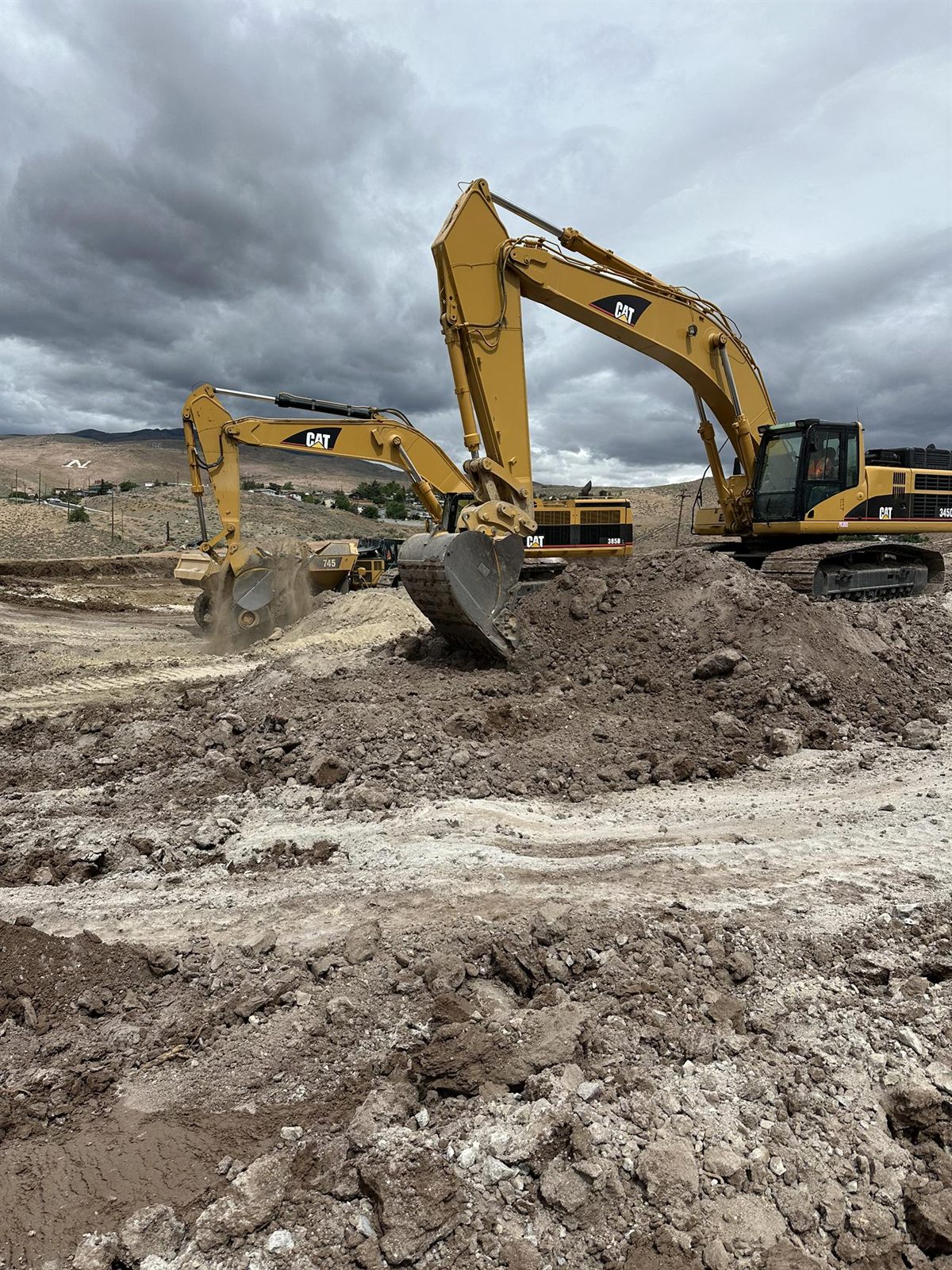 Two large Caterpillar excavators cutting into a bench together under a heavy storm sky, an articulated hauler waiting behind.